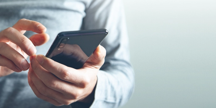 Nahaufnahme einer Person, die ein Smartphone in der Hand hält und den Touchscreen mit einer Hand bedient. Die Person trägt ein graues Langarmshirt, und der Hintergrund ist unscharf und hell gehalten