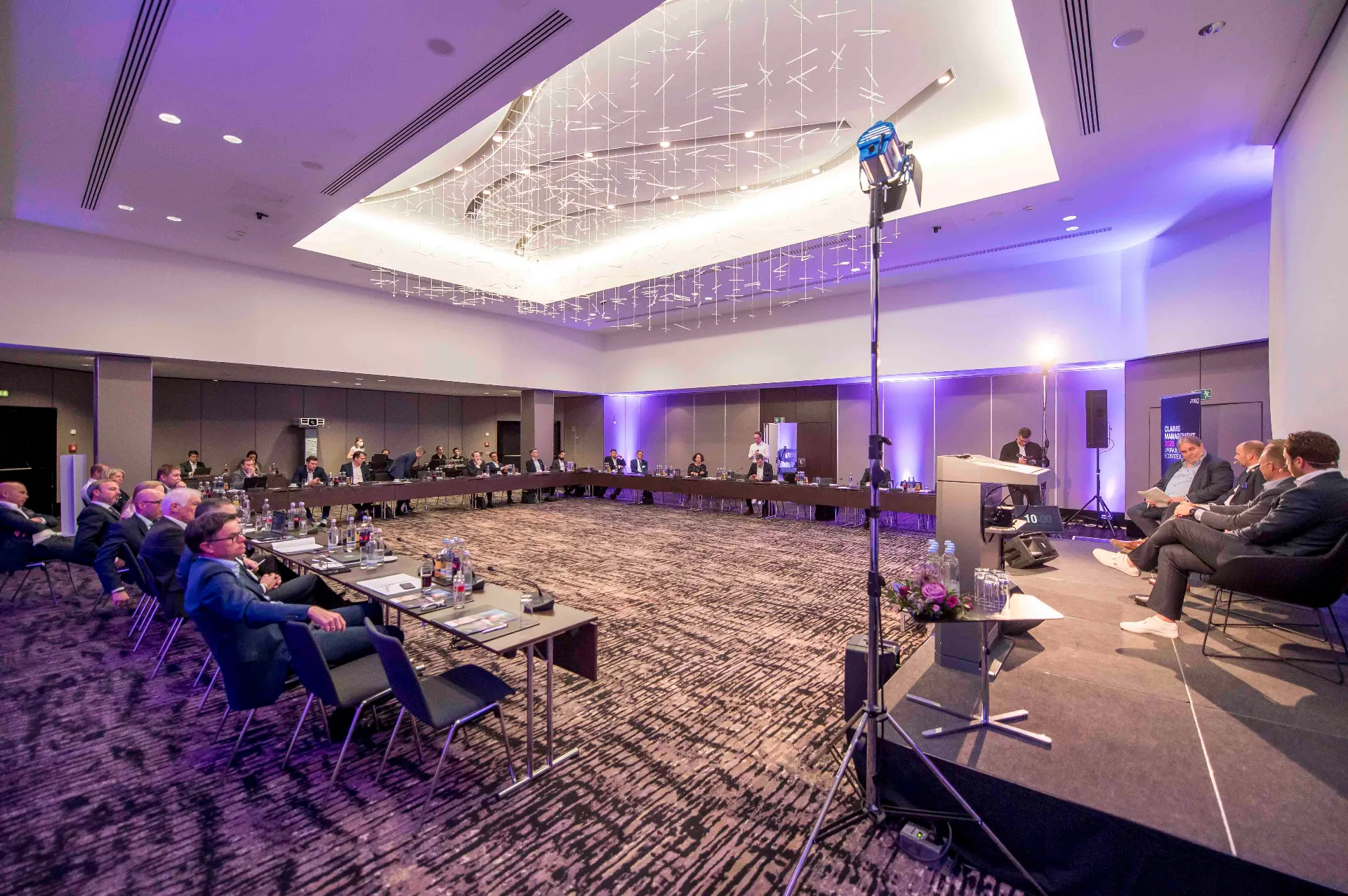 Zahlreiche Vertreter von Versicherungsunternehmen sitzen um ein Podest mit vier Herren versammelt und verfolgen die Diskussion. 